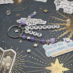 May include: A keychain spelling "LAVENDER HAZE" in white letter beads, with purple and clear beads, and silver star charms. The background is dark with gold starburst and star accents. A decorative accessory.