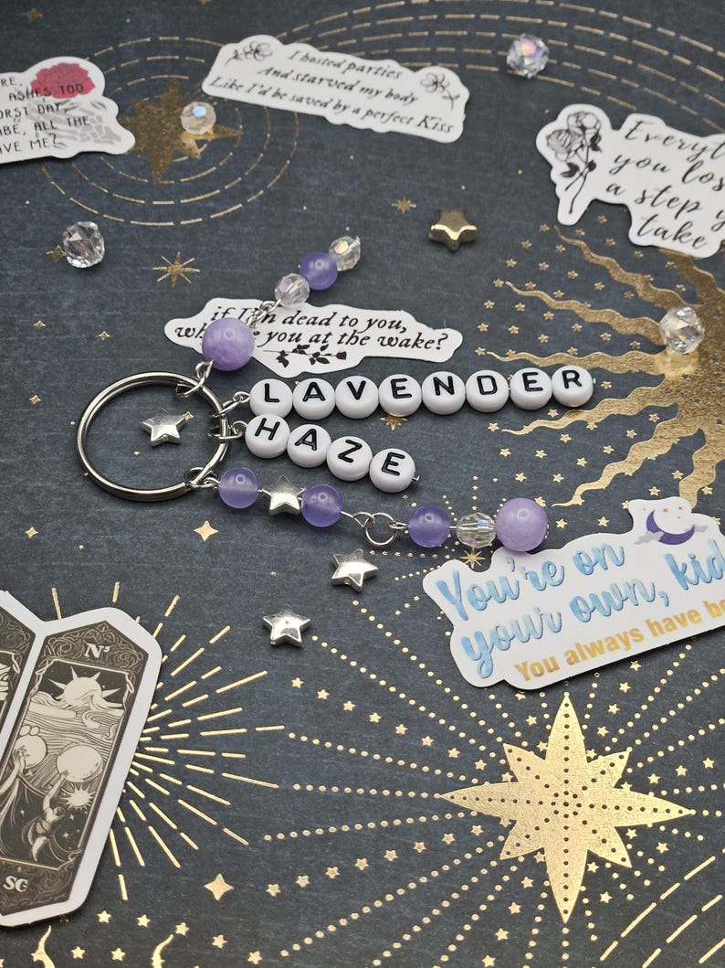 May include: A keychain spelling "LAVENDER HAZE" in white letter beads, with purple and clear beads, and silver star charms. The background is dark with gold starburst and star accents. A decorative accessory.