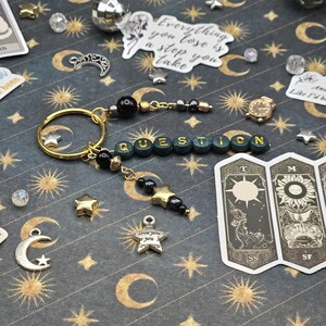 May include: A gold keyring with the word "QUESTION" spelled out in green and gold beads. The keyring features black beads, star charms, and a small gold watch charm. The background is a dark patterned surface with moons and stars.