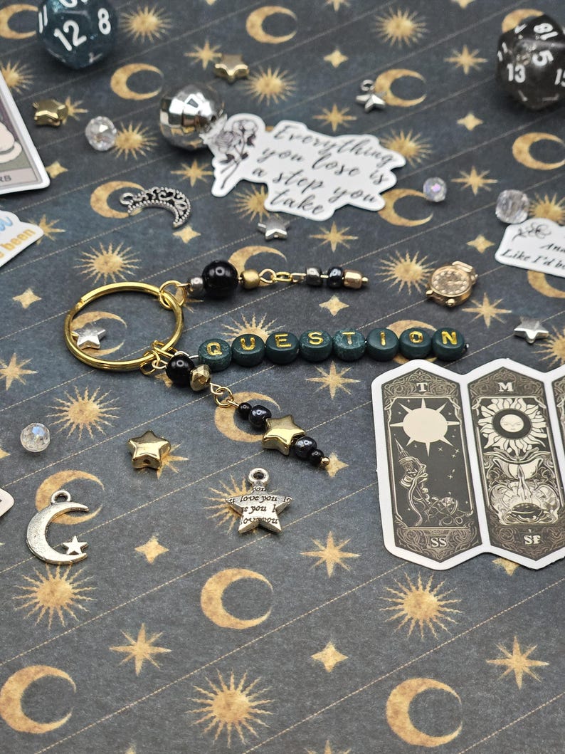 May include: A gold keyring with the word "QUESTION" spelled out in green and gold beads. The keyring features black beads, star charms, and a small gold watch charm. The background is a dark patterned surface with moons and stars.