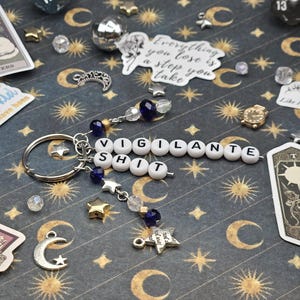 May include: A silver keyring with the words "VIGILANTE SHIT" spelled out in white letter beads. The keyring has star and bead charms in blue, gold, and clear. The background is a dark blue pattern with moons and stars.