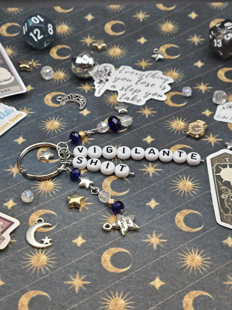 May include: A silver keyring with the words "VIGILANTE SHIT" spelled out in white letter beads. The keyring has star and bead charms in blue, gold, and clear. The background is a dark blue pattern with moons and stars.