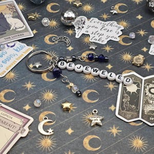 May include: A keychain spelling "BEJEWELED" in white letter beads, with blue beads and charms, including a star and a crescent moon. The background is a dark blue surface with gold celestial designs and tarot cards.