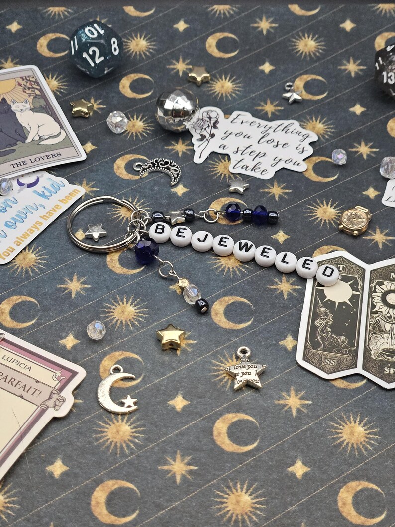 May include: A keychain spelling "BEJEWELED" in white letter beads, with blue beads and charms, including a star and a crescent moon. The background is a dark blue surface with gold celestial designs and tarot cards.