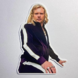May include: A sticker featuring a person with long blonde hair wearing a dark purple velvet tracksuit with white stripes. The person is wearing multiple rings and a gold necklace. The sticker is on a white background.