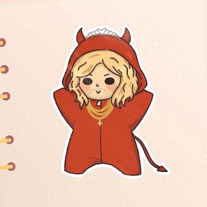 May include: A sticker featuring a cartoon character dressed in a red devil costume with a hood, horns, and a tail. The character has blonde hair, a beard, and a gold necklace with a cross pendant. A silver tiara is on the character's head.