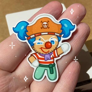 May include: A colorful sticker featuring a cartoon pirate clown character. The character has blue hair, an orange pirate hat with a skull, and a red and white striped shirt. The clown is winking and has a red nose.