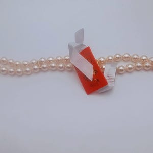 May include: A double-strand pearl necklace with a gold-coloured clasp. The pearls are a light cream colour. An orange tag with white ribbon is attached to the necklace.