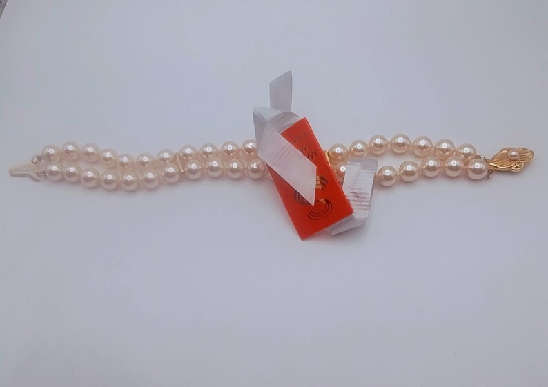 May include: A double-strand pearl necklace with a gold-coloured clasp. The pearls are a light cream colour. An orange tag with white ribbon is attached to the necklace.