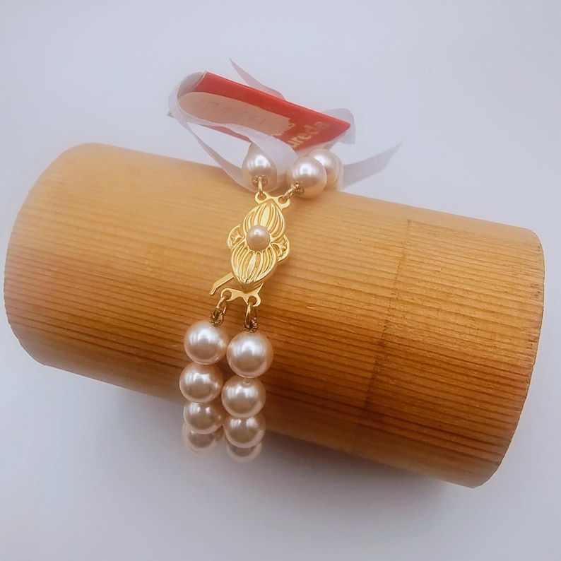 May include: A double-strand pearl bracelet with a gold clasp. The clasp has an ornate, floral design. The pearls are a creamy off-white colour. The bracelet is displayed on a light brown wooden cylinder, with a red tag attached.