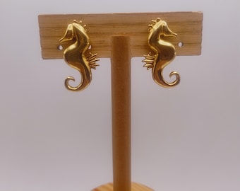 May include: A pair of gold-colored seahorse stud earrings. The earrings are displayed on a wooden jewelry stand. The seahorses have detailed features, including a curved body and a textured tail.