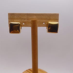 May include: A pair of gold-colored square stud earrings. The earrings are displayed on a wooden jewelry stand. The earrings have a smooth, reflective surface, and the square shape is slightly rounded at the edges.