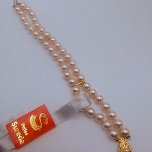 May include: A double-strand pearl necklace with a gold clasp and accents. The necklace features small, round, light-coloured pearls. A red tag with gold text that reads "Sureda Perlas" is attached.