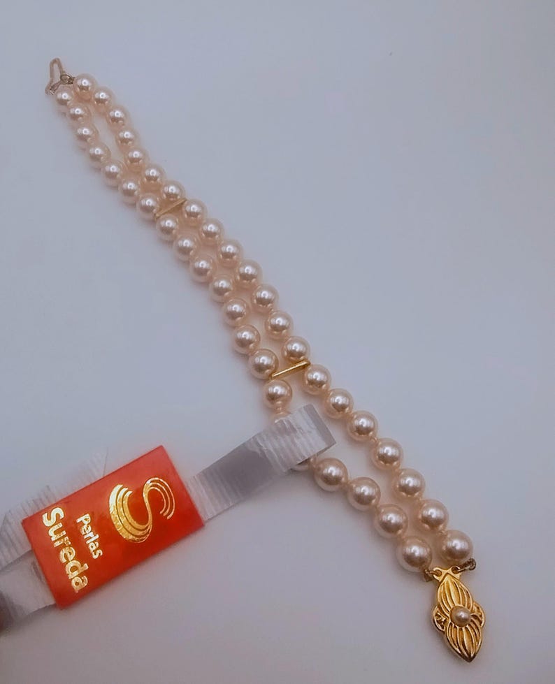 May include: A double-strand pearl necklace with a gold clasp and accents. The necklace features small, round, light-coloured pearls. A red tag with gold text that reads "Sureda Perlas" is attached.