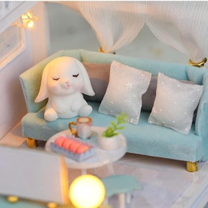 May include: Miniature dollhouse scene featuring a light blue sofa with white pillows and a white bunny figurine. A small table holds a cup, a plant, and a tray of treats. The scene is illuminated by a warm light.