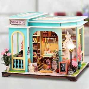 May include: A miniature tailor shop model with a teal and white exterior. The shop features a sign that reads "TAILOR SHOP". Inside, there are shelves, sewing supplies, and a mannequin. Pink flowers and a street lamp add decorative touches to the scene.