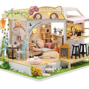 May include: A miniature cat cafe dollhouse with a charming design. Features a cafe area with a small chalkboard menu, seating, and a cat-themed interior. Includes a cat bed, cat trees, and decorative flowers. The sign says "Cat & Coffee" and "Lovely Cats Home".