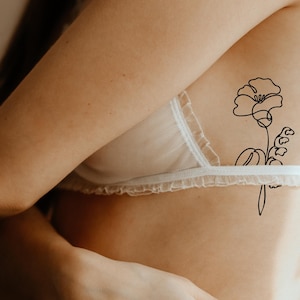 Op de afbeelding: Een zwarte lijntekening van een bloem en bladeren is op de huid getatoe&euml;erd. Het ontwerp bevindt zich aan de zijkant van de torso. Een witte, kanten beha is ook zichtbaar.