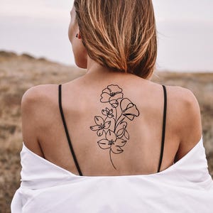 Op de afbeelding: Een zwart bloemen tattoo-ontwerp op de rug van een persoon. De tattoo toont een lijntekening van verschillende bloemen en stelen. De persoon draagt een wit overhemd en een zwarte top met spaghettibandjes.