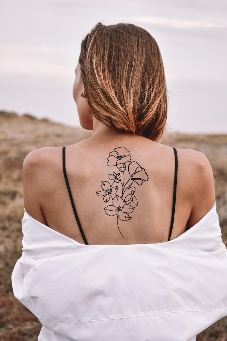 Op de afbeelding: Een zwart bloemen tattoo-ontwerp op de rug van een persoon. De tattoo toont een lijntekening van verschillende bloemen en stelen. De persoon draagt een wit overhemd en een zwarte top met spaghettibandjes.