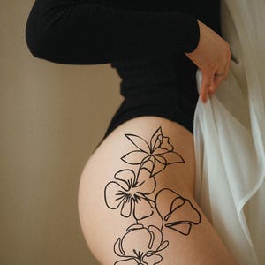 Op de afbeelding: Een zwarte bodysuit met lange mouwen. Een zwart bloemenlijnkunst tattoo-ontwerp is zichtbaar op de dij. Een witte, doorschijnende stof wordt in de hand gehouden.