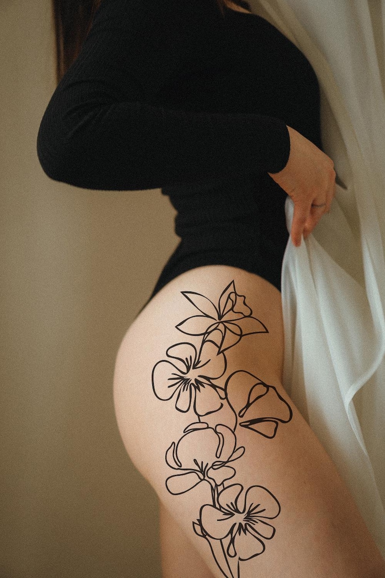 Op de afbeelding: Een zwarte bodysuit met lange mouwen. Een zwart bloemenlijnkunst tattoo-ontwerp is zichtbaar op de dij. Een witte, doorschijnende stof wordt in de hand gehouden.