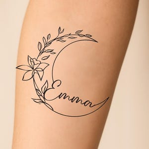 Può includere: Un tatuaggio a linea nera con un design a forma di mezzaluna con accenti floreali e di foglie. Il nome "Emma" è scritto in corsivo all'interno della curva della luna. Il disegno è su una carnagione chiara.