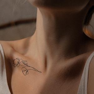 Op de afbeelding: Een minimalistisch zwart bloemen tattoo ontwerp op het sleutelbeen. De tattoo heeft een delicate omtrek van twee bloemen en een stengel. De persoon draagt een witte tanktop.