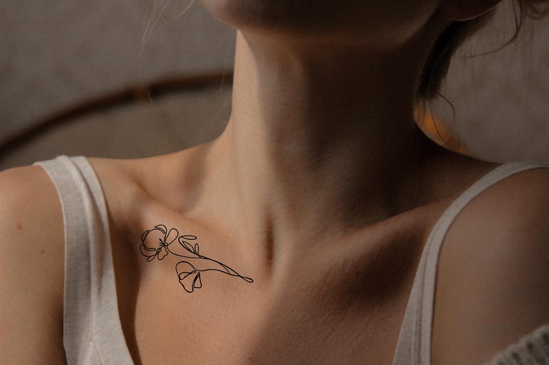 Op de afbeelding: Een minimalistisch zwart bloemen tattoo ontwerp op het sleutelbeen. De tattoo heeft een delicate omtrek van twee bloemen en een stengel. De persoon draagt een witte tanktop.