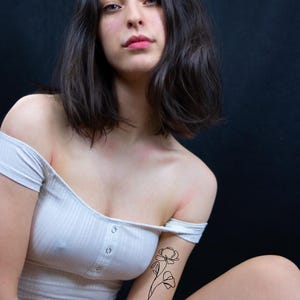 Op de afbeelding: Een persoon draagt een witte off-the-shoulder jurk met een bloementattoo op haar arm. De tatoeage is een eenvoudige lijntekening van een bloem. De persoon heeft donker haar en kijkt in de camera.