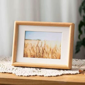 Op de afbeelding: Een rechthoekige houten fotolijst met een witte passe-partout, met een landschapsfoto van een tarweveld. De lijst staat op een houten oppervlak, met daaronder een wit kanten kleedje.