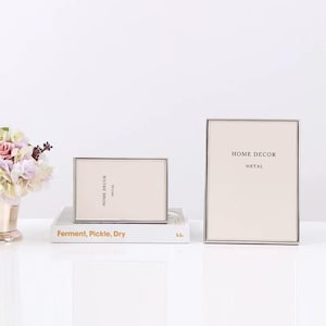 Puede incluir: Dos marcos de fotos rectangulares de metal con interior de color neutro. Los marcos están etiquetados como "HOME DECOR METAL". Un marco es horizontal, apoyado en un libro con el texto "Ferment, Pickle, Dry". El otro marco es vertical.