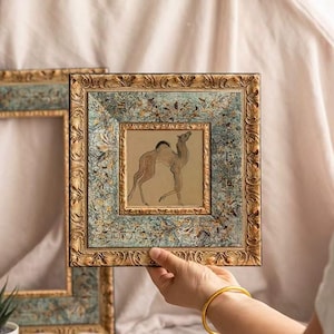 Pode incluir: Obra de arte emoldurada quadrada com uma ilustração de camelo. A moldura tem um acabamento dourado envelhecido com uma borda estampada em verde-azulado e dourado. O camelo é retratado em um estilo minimalista com tinta marrom e preta em um fundo bege.