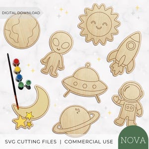 May include: A set of light-coloured wooden space-themed cutouts including a planet, alien, sun, rocket, UFO, moon with stars, and astronaut. A black paintbrush and a strip of five small paint pots are included. Text reads DIGITAL DOWNLOAD.