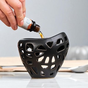 Pode incluir: Um difusor de óleos essenciais de cerâmica preta com um design vazado único. O óleo essencial está sendo derramado no difusor de uma garrafa. O difusor tem uma forma curva e está em uma superfície branca.