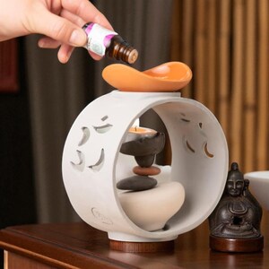 May include: A white ceramic essential oil diffuser with an orange dish on top. A hand is pouring oil from a small bottle. Inside the diffuser are stacked stones and a lit candle. A small statue sits to the side.