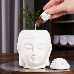 Pode incluir: Difusor de óleo de cabeça de Buda em cerâmica branca com tampa removível. Uma mão está derramando óleo essencial de um pequeno frasco no difusor. A cabeça de Buda tem olhos fechados e uma expressão serena. O fundo inclui uma prateleira de madeira e toalhas enroladas.