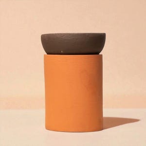 Peut inclure: Un pot de fleurs en céramique en deux parties au design moderne. La base est de forme cylindrique, de couleur orange vif, supportant un bol peu profond brun foncé. Convient aux petites plantes ou succulentes.