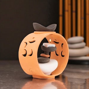 May include: Orange ceramic candle holder with a curved design and cut-out accents. A lit candle sits inside, with stacked stones and a white heart-shaped base. A dark grey decorative piece sits on top. The background features a stack of stones and bamboo.