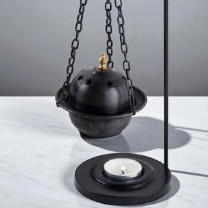 May include: A black metal incense burner with a hanging bowl-shaped top and a base with a lit tea light candle. The bowl has star-shaped cutouts and is suspended by chains. The top has a gold-colored finial.