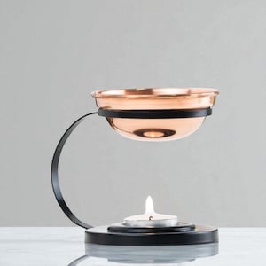 Puede incluir: Un difusor de aceites esenciales de color cobre con una estructura de metal negro. El difusor tiene un cuenco pequeño para el aceite y un portavelas debajo. La vela está encendida y el difusor está sobre una superficie blanca.