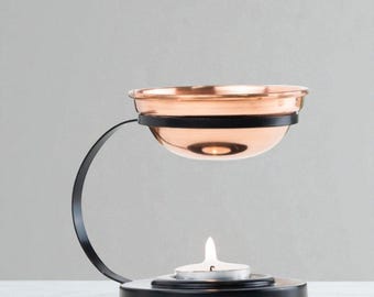 May include: A copper-colored essential oil diffuser with a black metal frame. The diffuser has a small bowl for oil and a candle holder below. The candle is lit, and the diffuser is on a white surface.