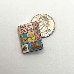 May include: A miniature "Betty Crocker's New Picture Cook Book" with colourful food illustrations. The small book is next to a British coin for size comparison. The book's cover features the title and images of a cake, a turkey, and other dishes.