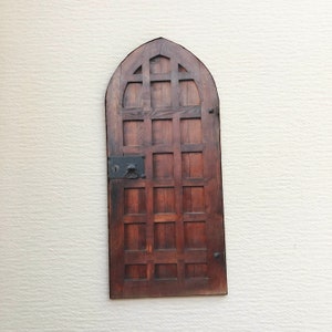 May include: A small, arched wooden door with a dark brown finish. The door features a grid-like design and a black metal lock with a handle. The door is mounted on a textured, off-white wall.