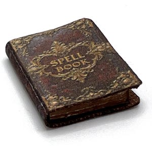 May include: A miniature, antique-style spell book with a dark brown cover and gold-colored ornate detailing. The words "SPELL BOOK" are embossed on the cover. The book's pages are visible, suggesting it is open.