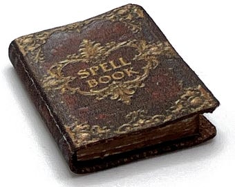 May include: A miniature, antique-style spell book with a dark brown cover and gold-colored ornate detailing. The words "SPELL BOOK" are embossed on the cover. The book's pages are visible, suggesting it is open.