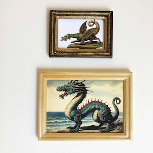 May include: Two framed dragon art prints. The top frame has a gold finish and contains a green and brown dragon illustration. The bottom frame has a light wood finish and features a blue and red dragon illustration with a seascape background.