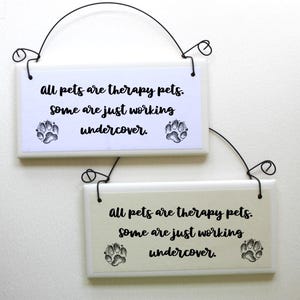 Peut inclure: Deux pancartes en céramique blanche avec du texte noir et des empreintes de pattes qui disent "Tous les animaux de compagnie sont des animaux de compagnie thérapeutiques. Certains travaillent juste sous couverture."