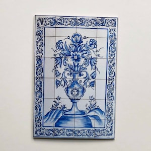 May include: A blue and white ceramic tile with a decorative design of a vase with flowers. The tile is surrounded by a blue and white border.