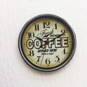 May include: A round, black framed clock with a white face and black hands. The clock face has the words "Fresh Brewed Coffee" and "Served Here Have a cup!" printed on it.